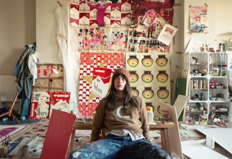 The Charlotte Rose art studio is shown full of new Pop art artworks in progress, with the artist sitting in the middle.
