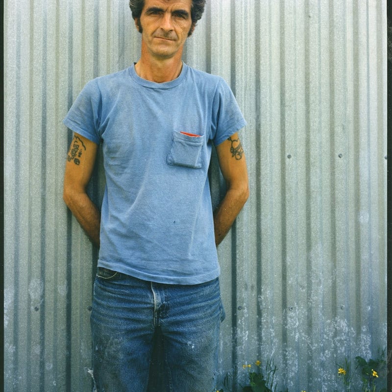 Bruce Wrighton Man with tattoed arms, in blue t-shirt, in front of green corrugated fence, yellow flowers at bottom Tirage C-print d'époque 20 x 25 cm Dim. papier: 20 x 25 cm