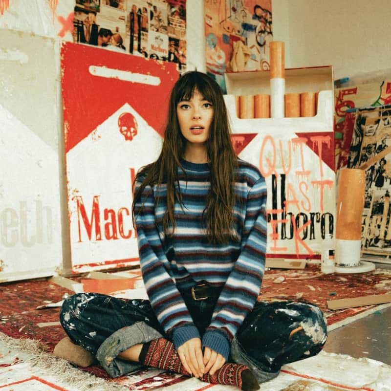 Charlotte Rose seated cross-legged on a paint-splattered rug, surrounded by bold, cigarette-themed art pieces, capturing the artist in her vividly expressive studio space
