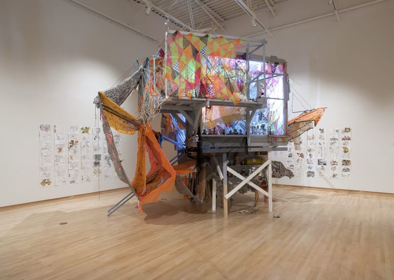 GREG SMITH TINA, 2018 Concrete, acrylic, polyester thread, cotton rope, cardboard, paper, graphite, colored pencil, manila folders, plaster, canvas, desk, door, chair, conveyor belt, conveyor drums, florescent lights, latex paint, newspaper, nylon fabric, parachute, plastic sheeting, rattan, wire, electronics, hardware, wood, salt and pepper shakers, dishes, lapel pins, plant and grow light 156 x 195 x 125 in.