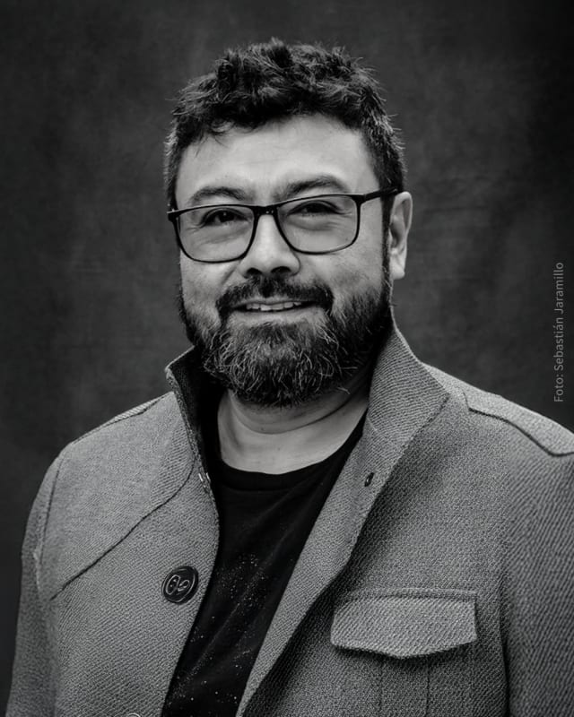 Portrait of Colombian artist Miler Lagos, photographed in black and white by Sebastián Jaramillo. The image shows him in a textured jacket and glasses, gazing confidently at the camera against a neutral background, reflecting his thoughtful and experiment