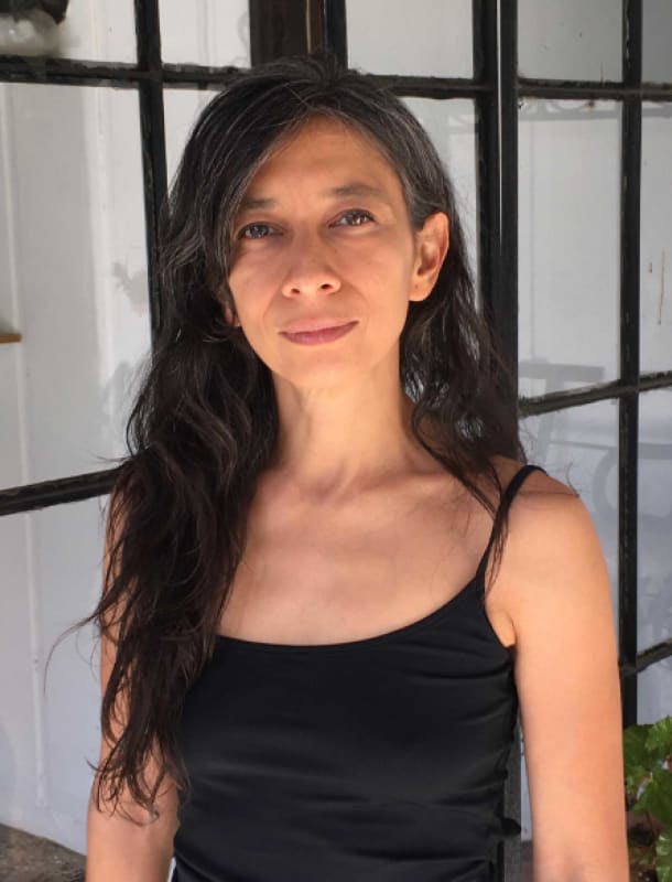 Portrait of Regina José Galindo standing in front of a window with black metal frames, wearing a black top. Her expression is calm and introspective, her long dark hair falling over one shoulder. The image conveys the quiet strength and presence character