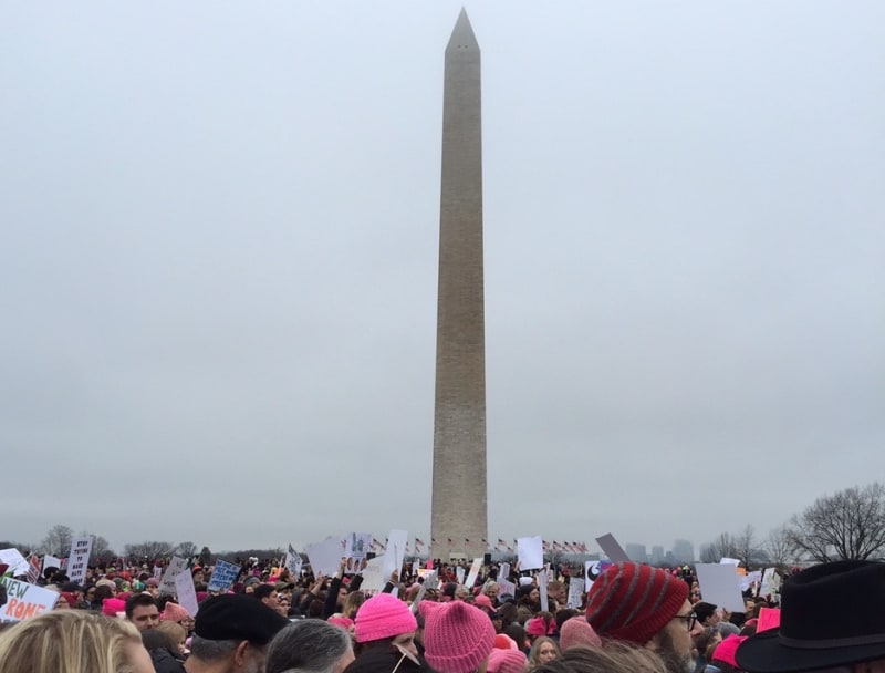 The Washington Monument is seen in the background as a large crowd of people, many wearing pink "pussy" hats, march in Washington