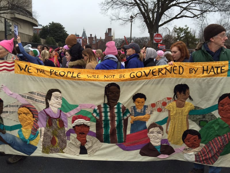 Protestors at the Women's March hold up a banner with the words "we the people" along the top, and representations of different famous Americans below it