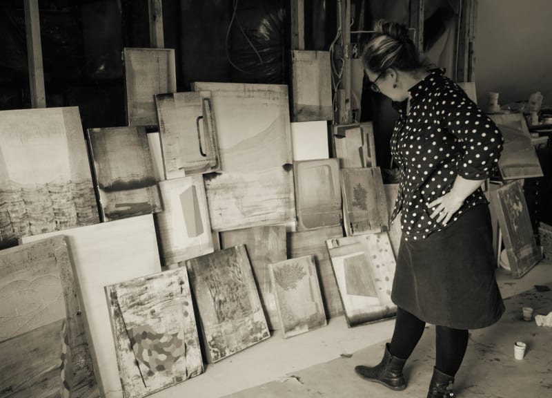 A black and white photo of Gabe Brown in her studio standing and looking at stacks of her paintings against the wall