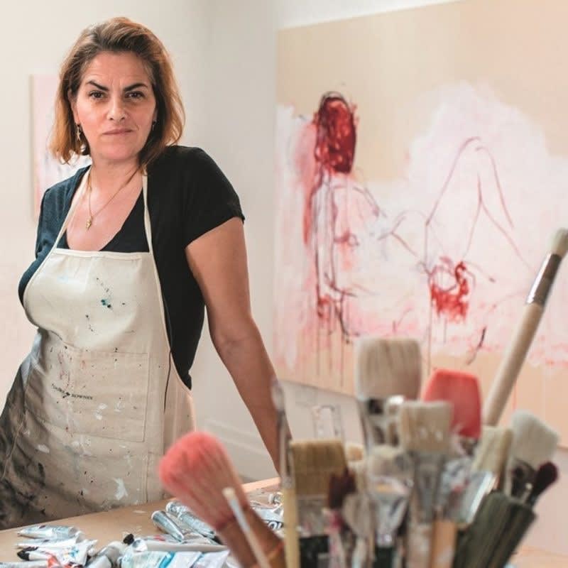 Tracey Emin standing in her art studio with painting supplies and a canvas featuring a red abstract figure in the background.