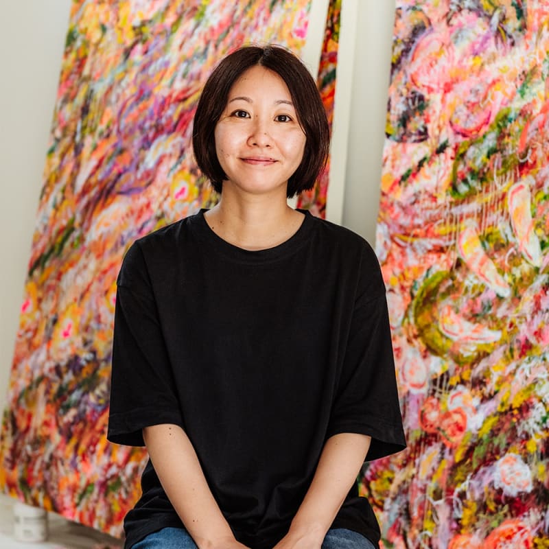 Artist Ayako Rokkaku sitting in front of her vibrant abstract paintings
