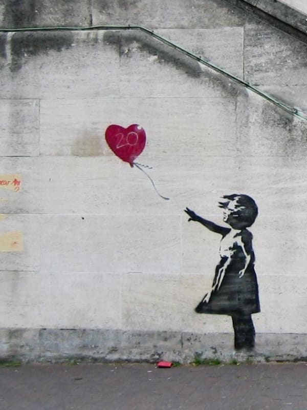 Street art by Banksy titled 'Girl With Red Balloon' depicting a young girl reaching for a heart-shaped red balloon on a concrete wall.