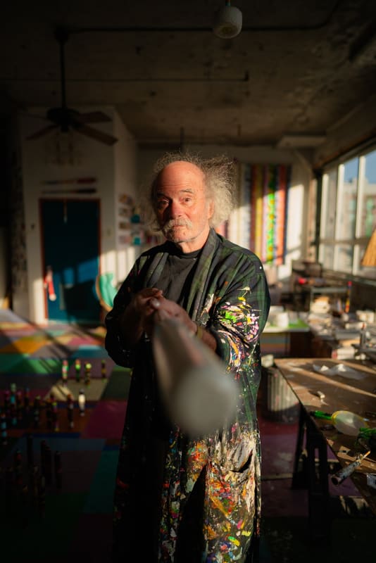 An artist, Steve Silver, holding out a cane amidst sculptures and paintings in an industrial loft setting flooded with late afternoon light