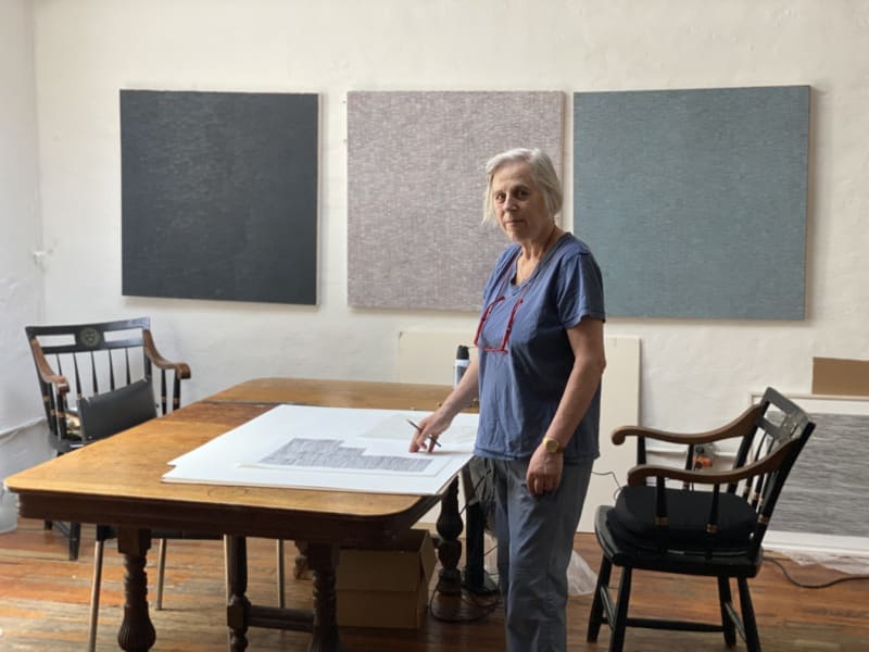 Artist Inger Johanne Grytting standing in her NYC studio with an unfinished drawing on a light wood table and three minimalist paintings hanging on a white brick wall, 2022