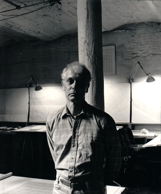 Artist Will Insley stands in front of a column in his NYC Bowery studio with architectural drawings in the background in 1983