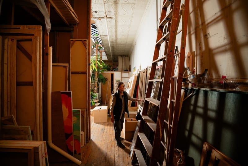 Photograph of an artist standing amongst canvases in a loft setting