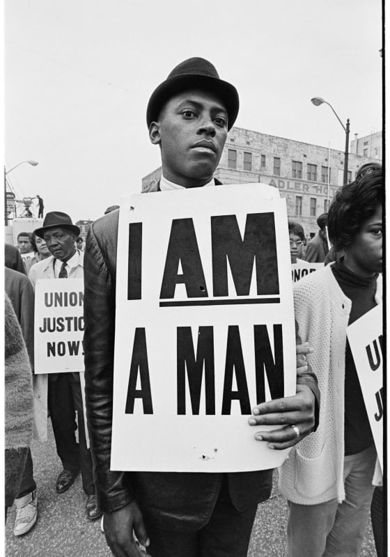 Black & white photograph of African-American man carrying a board with the words “I Am A Man”