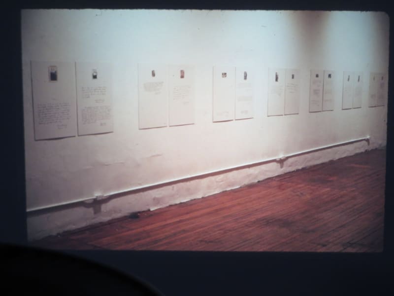 Seven pairs of white prints with black text, all identical in size and shape, hang on a white wall in a gallery setting.