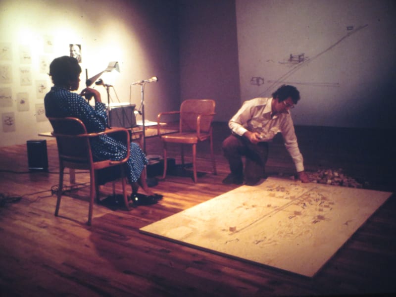 A man leans over a canvas placed on a wood floor. A woman sits in a chair on his left in front of speakers and a microphone. A collection of small black and white prints hangs on the wall behind them.