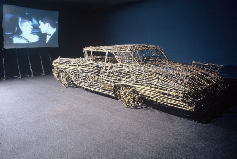 A 1958 Cadillac Eldorado Biarritz is recreated from twigs and bamboo and placed in front of a movie screen. The walls are painted dark blue and the carpet is gray to look like a drive in theater.