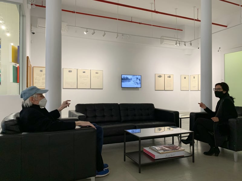 A man and a woman sit across from each other on black couches surrounding a coffee table in a gallery setting. On the wall behind them are 9 small white framed prints and a television.