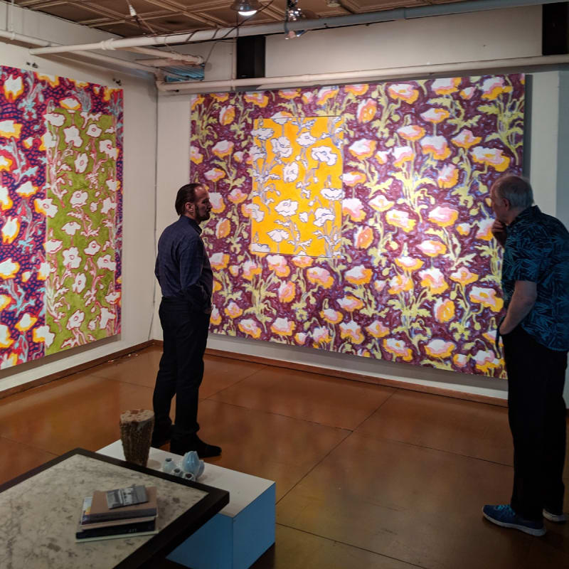 Two paintings hanging on white walls in a studio setting. Two gentlemen stand on either side of the right-hand painting viewing and discussing the piece.