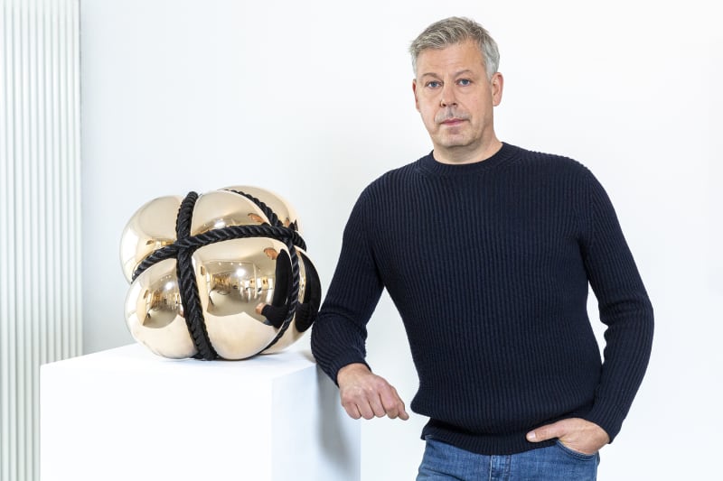 Portrait of Stephan Marienfeld alongside his sculpture “Bondage,” featuring a reflective golden surface bound with black rope. Copyright © Buysse Gallery, Studio Stephan Marienfeld.