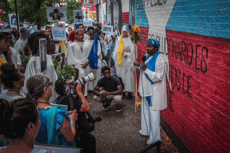 Campos-Pons addresses the procession as it stops at the Dos Alas (“Two Wings”) mural on 105th Street and Third Avenue. Credit: Graham Dickie/The New York Times