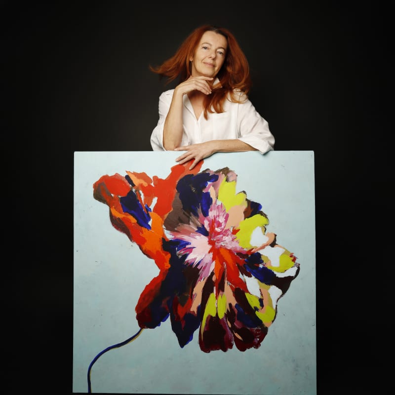 Portrait of artist Caroline Muscat standing behind one of her paintings, featuring a bold, expressive flower form in vivid reds, blues, yellows and pinks against a pale blue background.