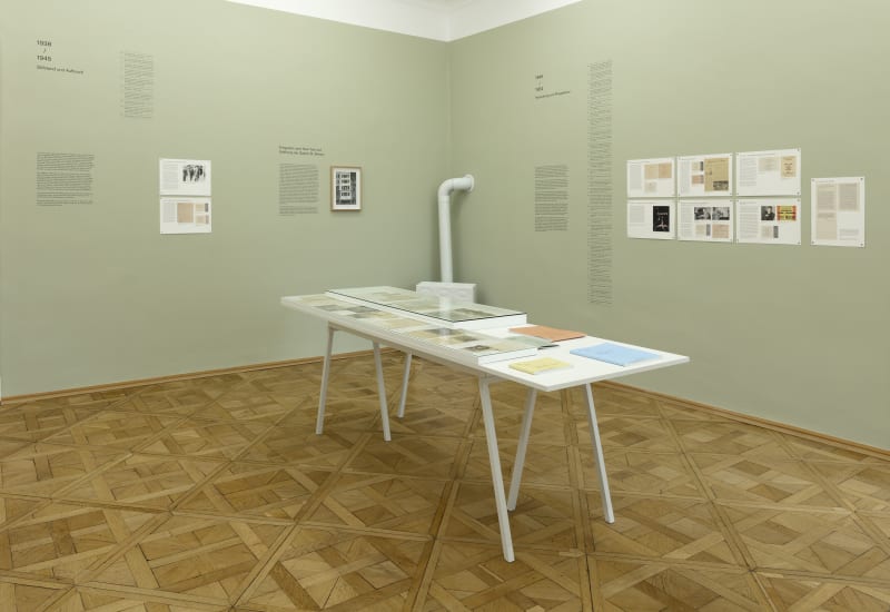 Interior view of an exhibition gallery displaying documentary papers on a central table with descriptive signage and texts on a sage green walls. the parquet floors are a medium wood tone.
