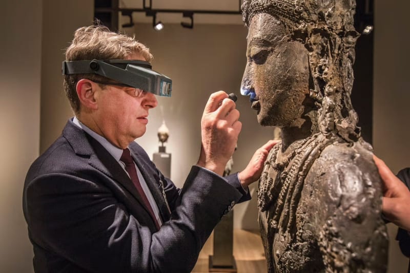 A brown haired white male in a suit wears magnifier glasses as he holds a flashlight up to inspect a sculpture bust of a human figure at an art fair