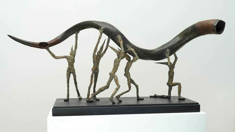 Bronze sculpture of elongated human figures carrying a large curved horn, arranged in a dynamic procession on a black base.
