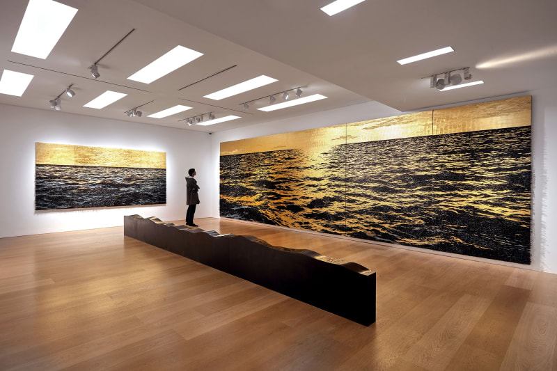 Installation view of Yoan Capote's exhibition in a white walled gallery featuring black and gold paintings and a long low black stone sculpture