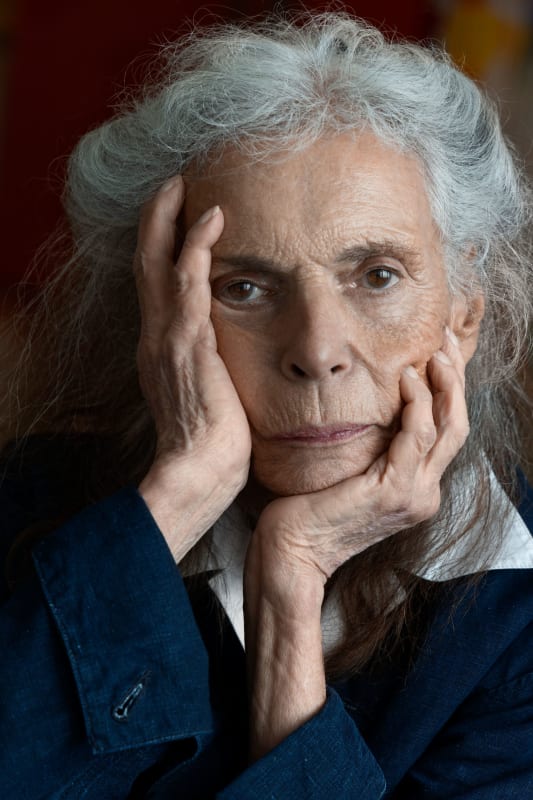Color image of Pat Steir looking directly into the camera with one hand underneath her chin and the other flat against the right side of her face.