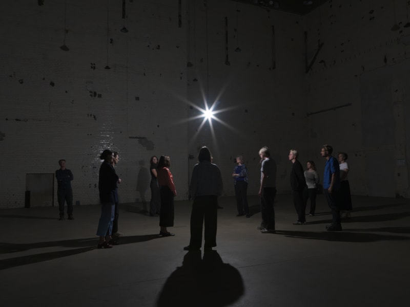 Cornelia Parker's installation at KINDL. An image of a dark warehouse space, a number of people gather around a single lightbulb, dramatic shadows are cast on the floor.