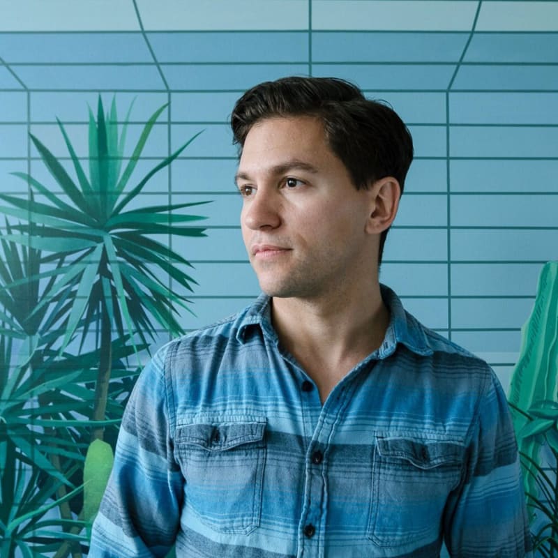 portrait of artist Robert Minervini - man in blue striped button down shirt looking off to side; blue painting in background with stripes and plant