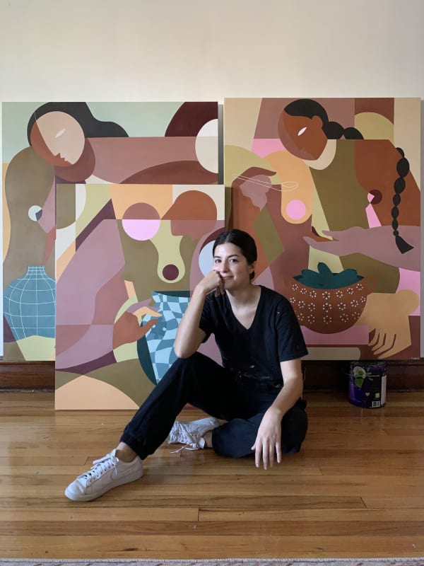 Liz Flores, a young woman, sits in front of her brown, pink, and other neutral tone paintings in her hardwood studio.