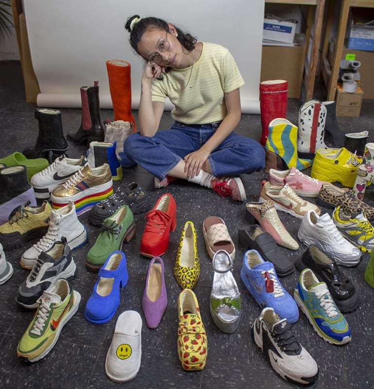 portrait of artist Didi Rojas (woman sitting on floor surrounded by her ceramic shoe sculptures)