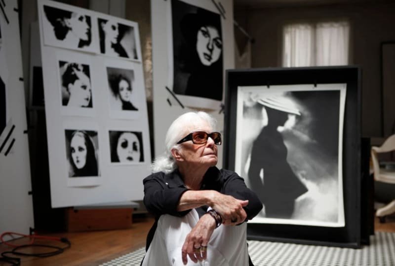 Lillian Bassman sitting on floor of her studio in sunglasses with her photographs behind her