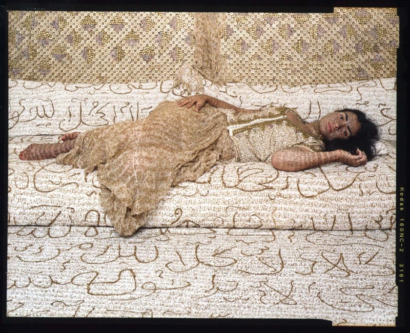 Woman reclining on daybed in Orientalist pose, her skin inscribed in Arabic calligraphy in henna, with golden bullets shimmering as luxurious background, by Lalla Essaydi