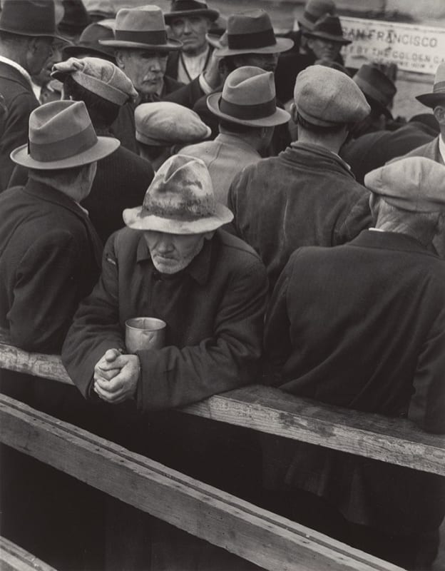 A black-and-white photograph by Dorothea Lange showing a group of men wearing hats and coats tightly gathered together, likely during the Great Depression. One elderly man stands at the front, leaning on a wooden railing with a weary expression, holding a