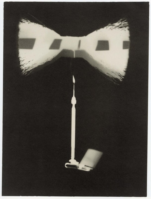Black-and-white rayograph by Man Ray showing silhouetted objects, including a bow-shaped form and elongated rod, against a dark background.