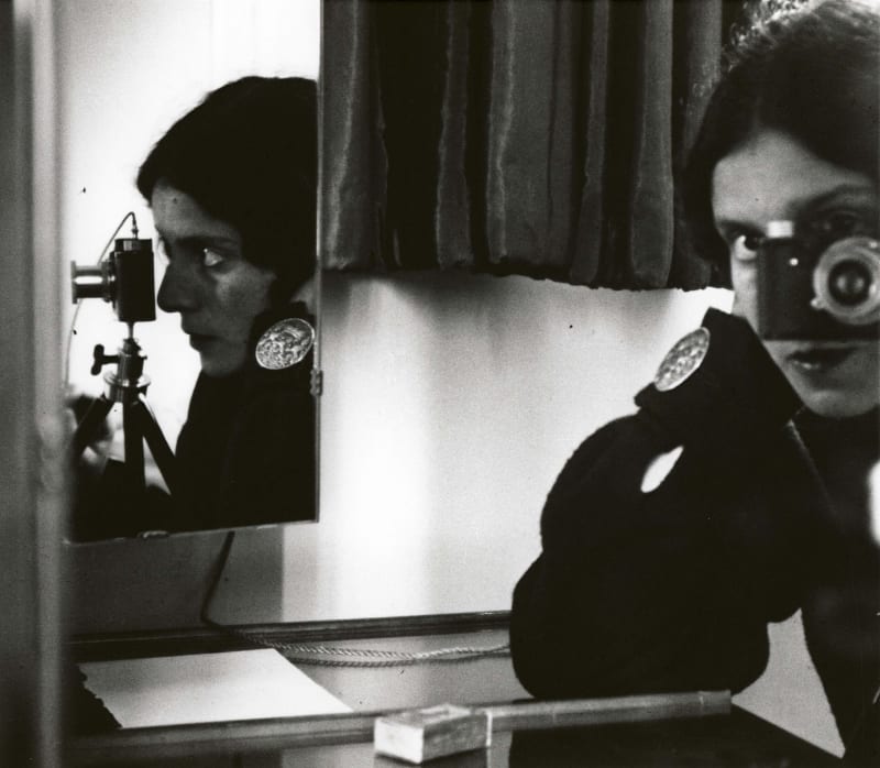 Black-and-white Ilse Bing self-portrait showing the photographer reflected in a mirror, Leica camera raised, with split reflections framing her face.