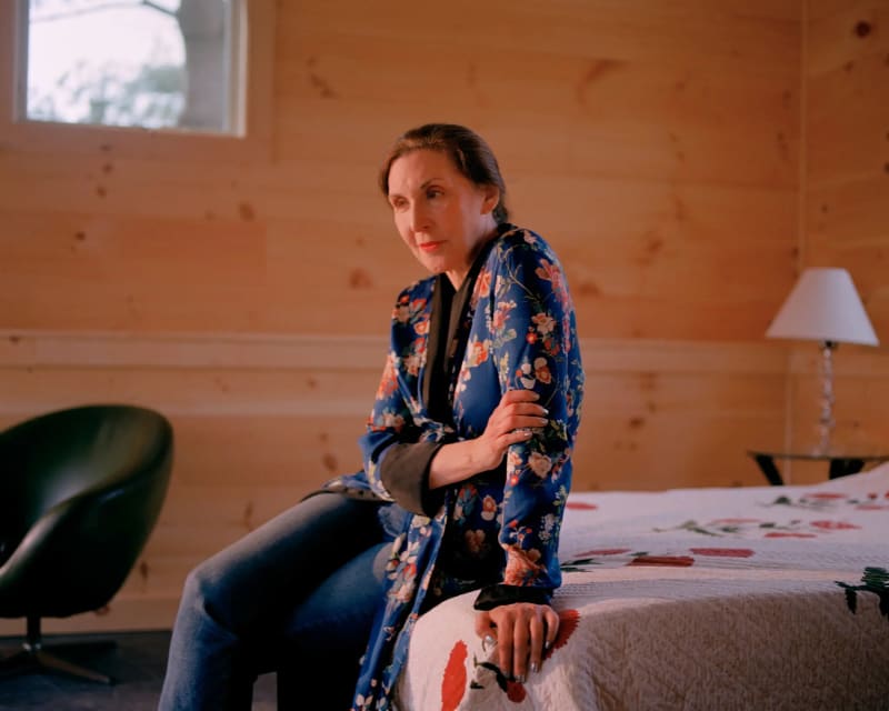 Portrait of Laurie Simmons wearing a floral blue robe and jeans, sitting thoughtfully on the edge of a bed in a warmly lit wooden room.