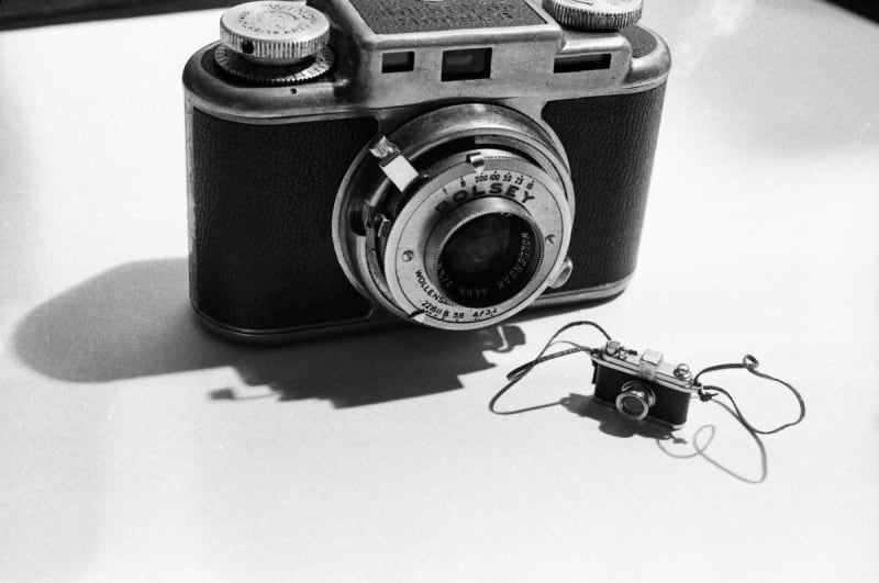 Black-and-white photograph by Laurie Simmons featuring a large vintage camera next to a tiny toy camera with a strap, casting shadows on a white surface.