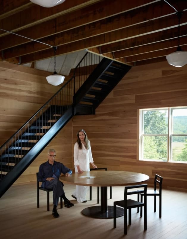 Two people pose in a wood-paneled room with a round table and modern staircase, seated and standing beneath exposed beams and soft natural light.