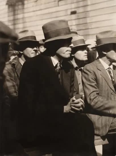 Dorothea Lange image of a group of men wearing suits and wide-brimmed hats sit closely together, facing forward in an outdoor setting with sunlight casting shadows