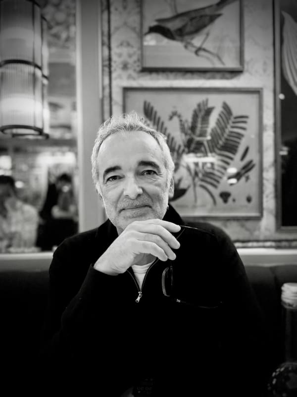 Black-and-white portrait of Joe Madeira seated in a café, holding his glasses with a relaxed expression, with botanical and bird illustrations framed on the wall behind him.