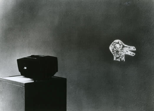 Grainy black and white image of a slide projector on a pedestal casting the image of an illustrated duck head.