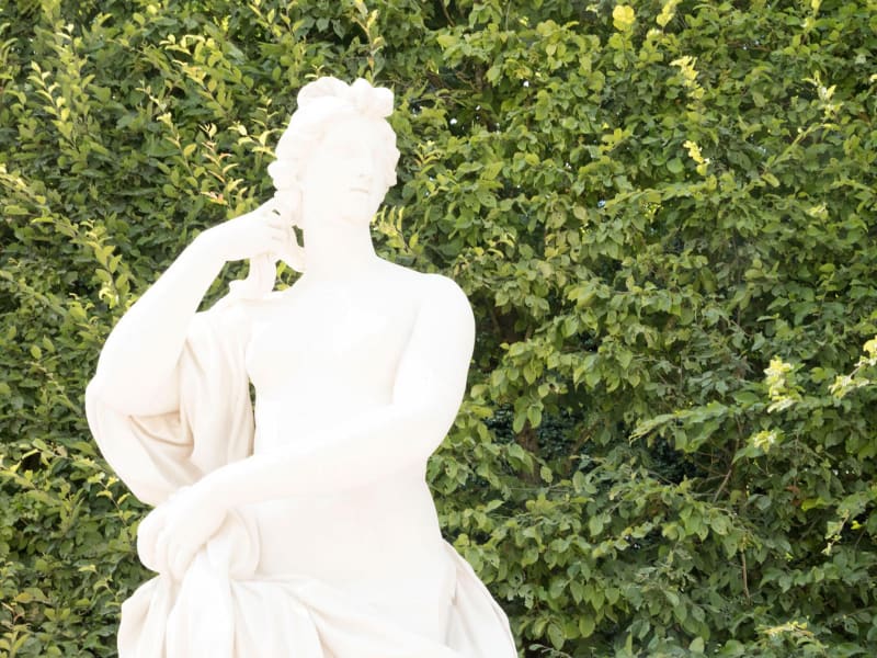 A white sculpture of a naked woman sitting with one arm resting on her right knee in front of greenery.