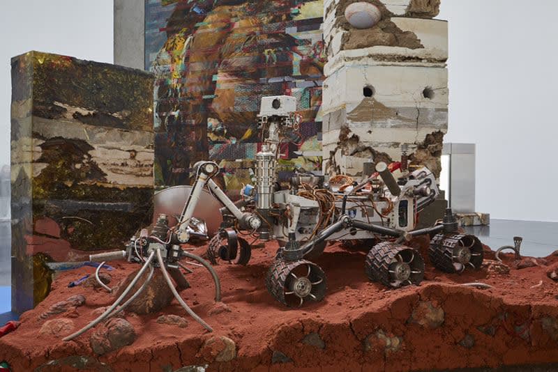 Installation of robotic sculpture mounted on red turf-like material. There is a camera on the robot.