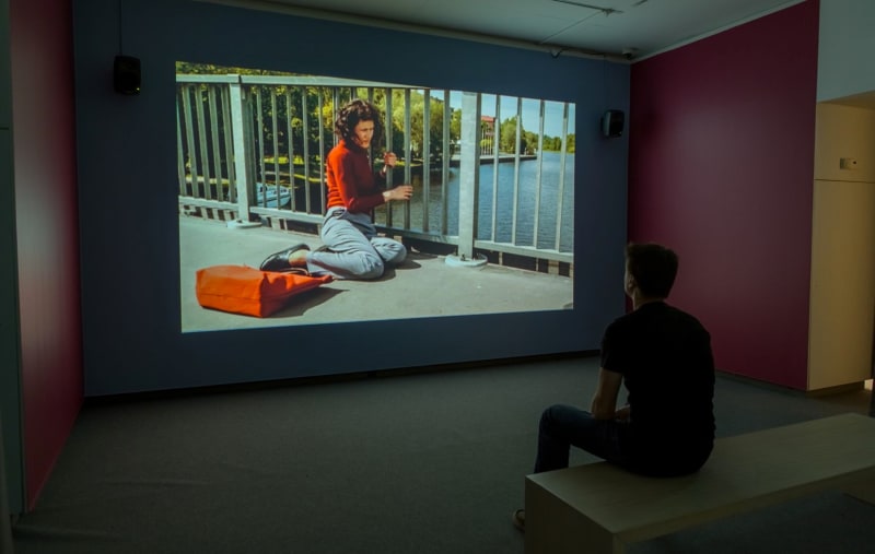 Gallery view of video still. In video, a person sits on a bridge, clutching the railing. Bag is strewn on the ground. A person watches the video.