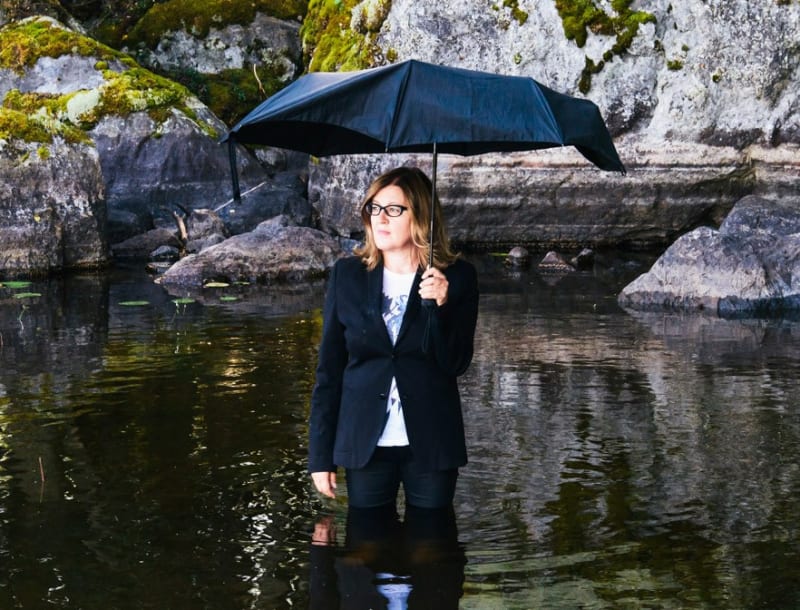A woman with an umbrella in a lake surrounded by moss and rocks.