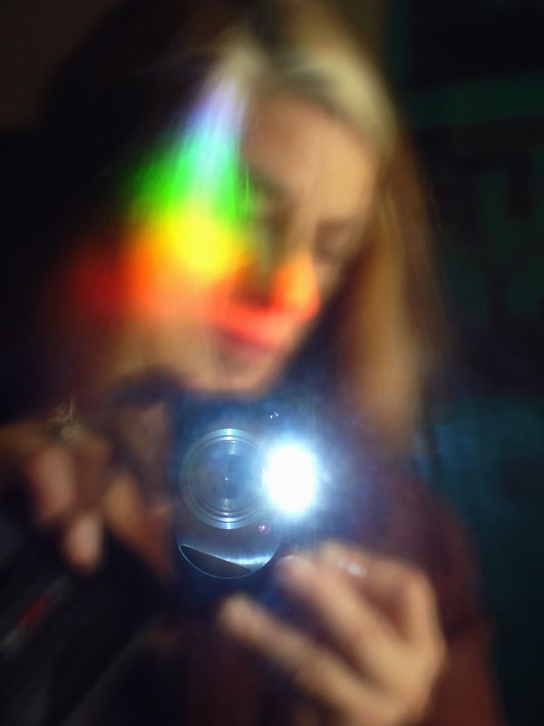 artistic photograph of person taking their own photo in a mirror. a rainbow light flare obstructs the face.